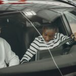 כמה מרוויח בוחן נהיגה - Black woman and caucasian man sitting inside a vehicle, discussing driving.